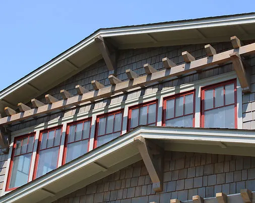 A close-up of a house with dark shingles, red-framed windows, and exposed wooden beams under a clear blue sky.
