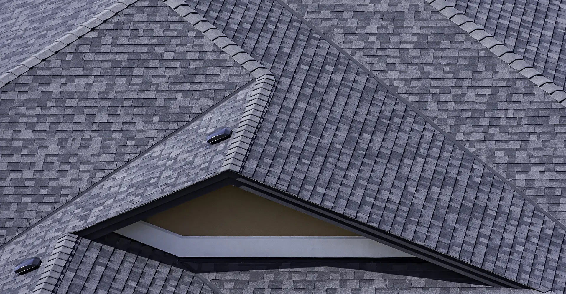 A close-up view of a gray asphalt shingle roof showing intersecting gables, roof vents, and textured shingles.