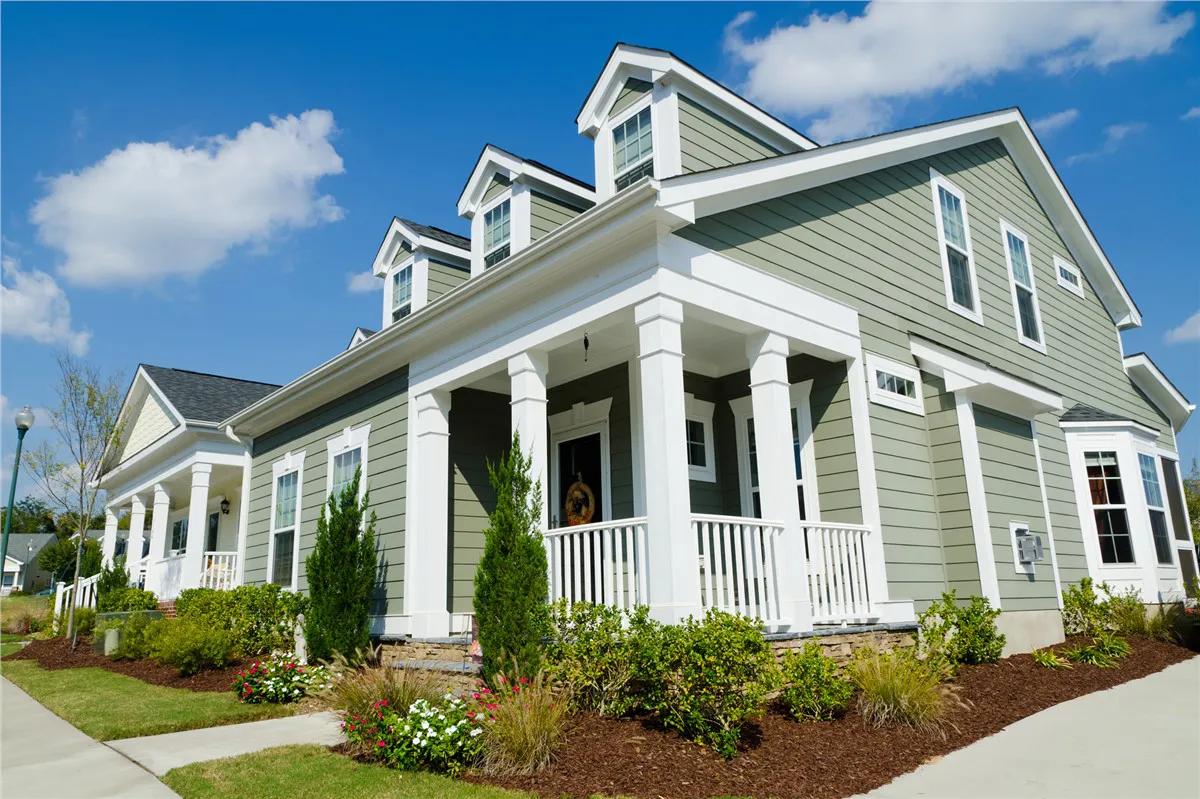 A light green colonial-style house with white trim and a front porch, set against a bright blue sky with scattered clouds.