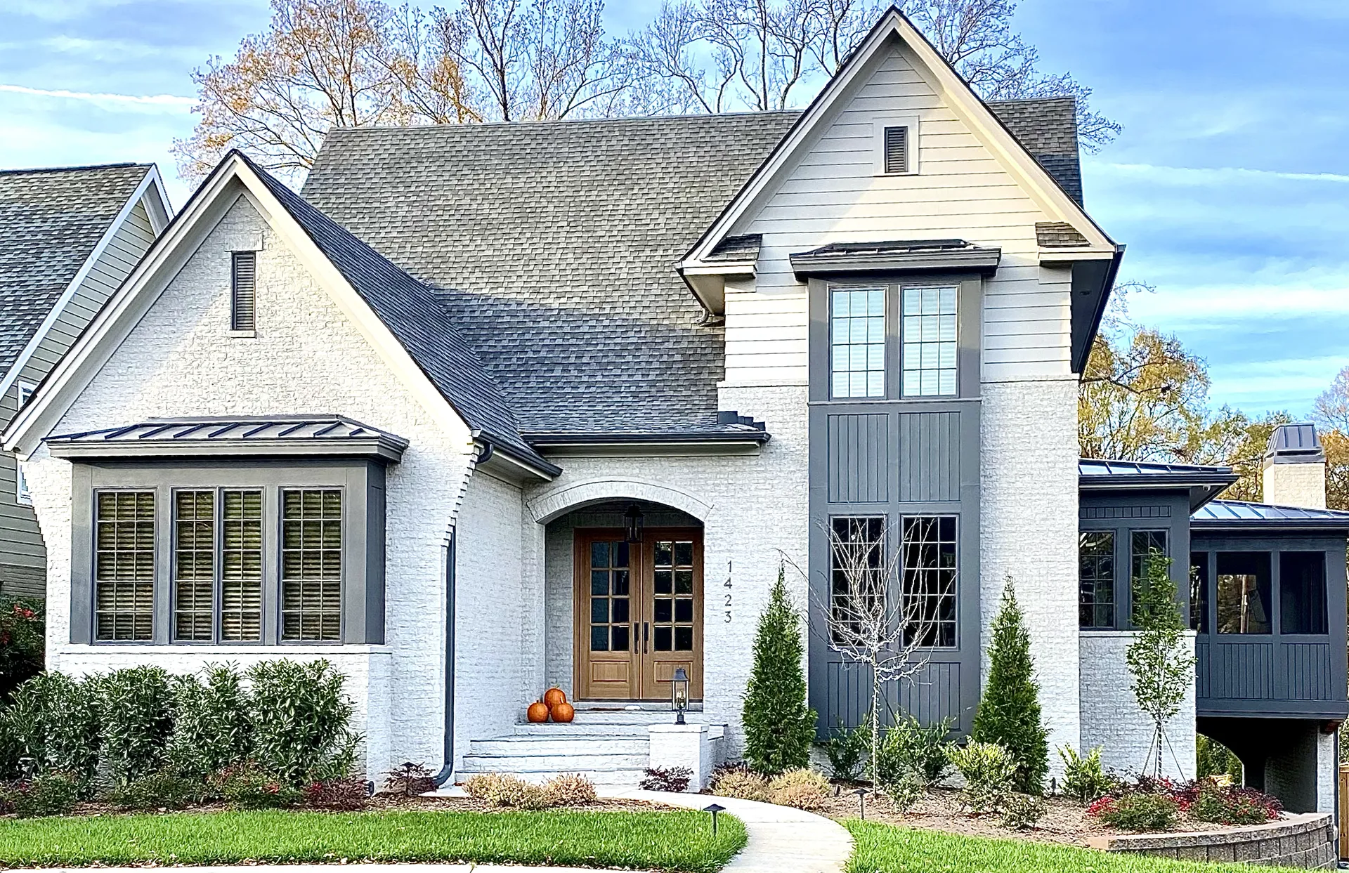 A stylish two-story home with white stucco and gray accents, featuring a wooden front door, pumpkins on the porch, and a well-manicured lawn.