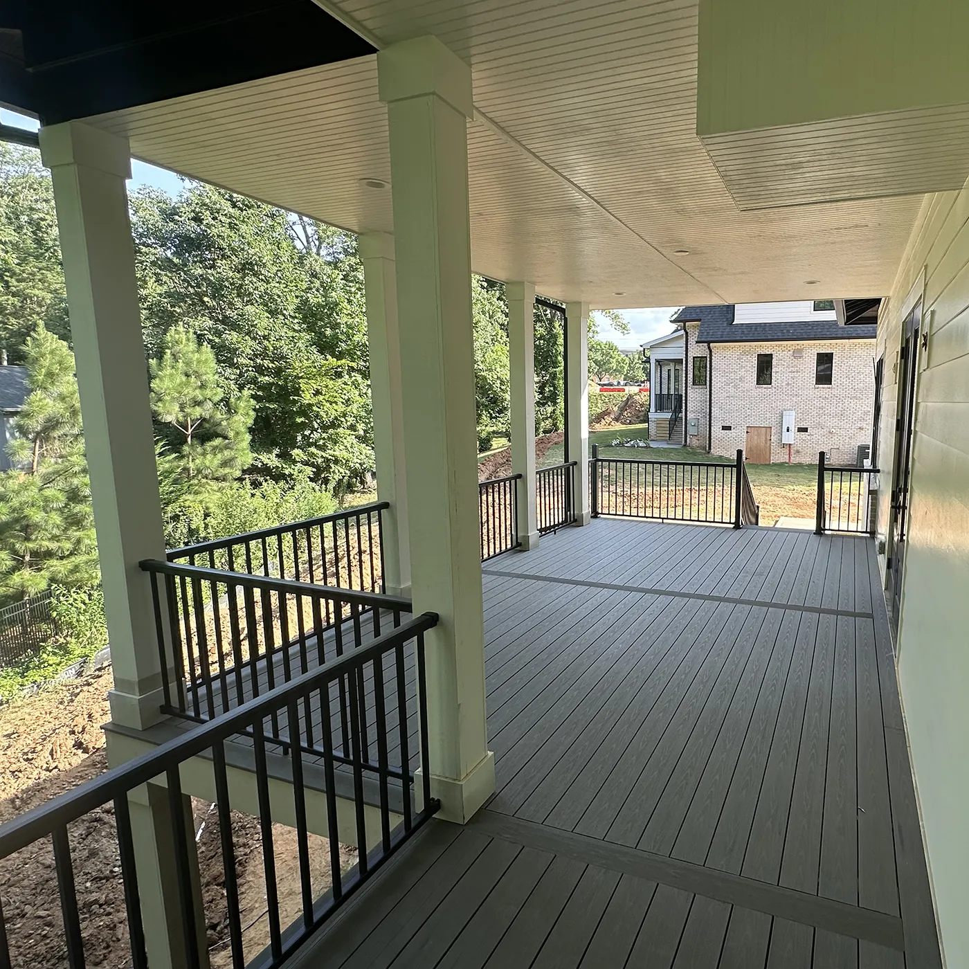 A spacious, newly constructed porch with gray decking and black railings under a light green roof, overlooking a wooded area and neighboring house.